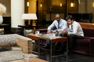 Two men in business attire sit on a sofa in a hotel setting, reviewing documents on a table with notebooks, glasses of water, and a calculator.