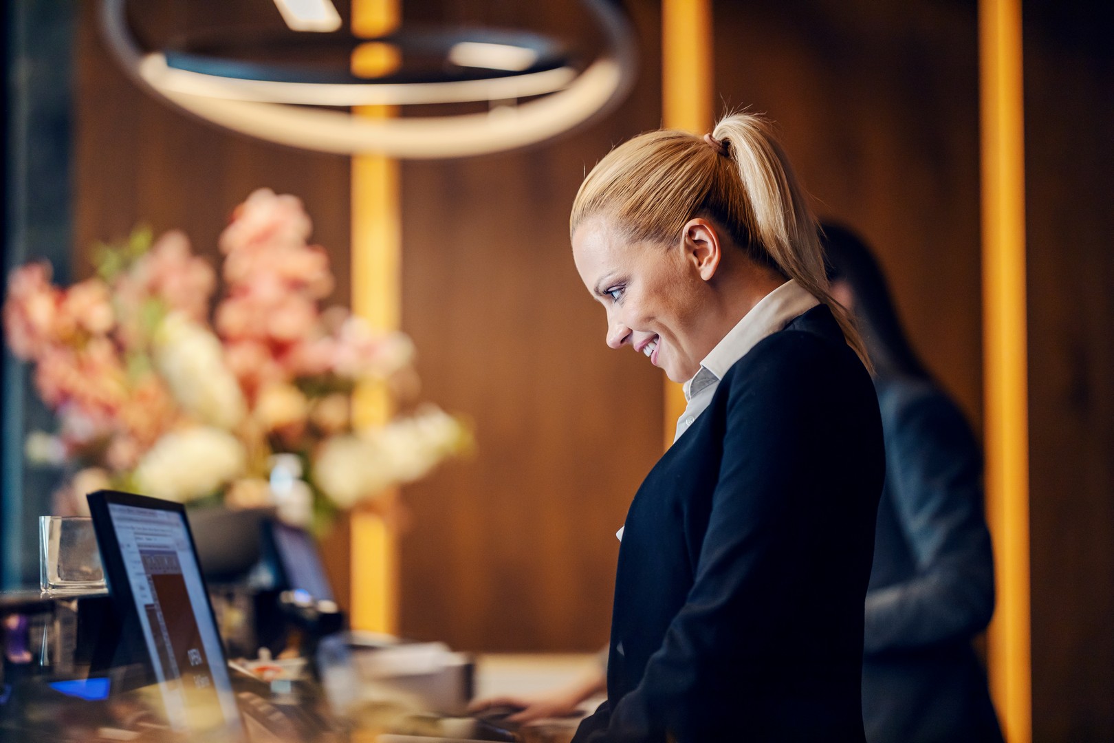 A woman with blonde hair in a ponytail smiling while working on a computer at a hotel reception desk, with a blurred person and flower arrangement in the background.